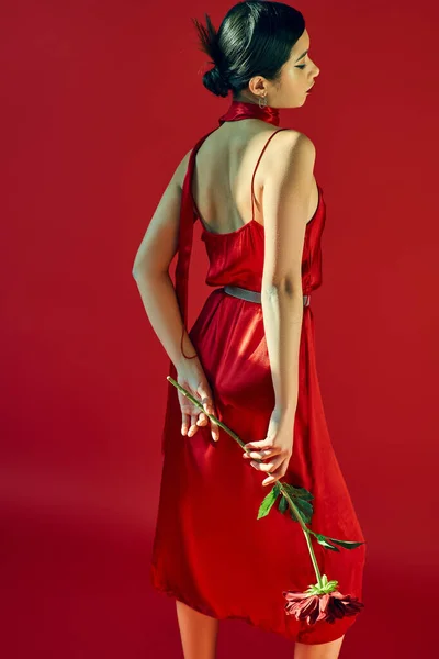 Mujer asiática joven y elegante con cabello moreno y peinado de moda de pie en pose elegante con peonía borgoña sobre fondo rojo, primavera de moda, generación z - foto de stock