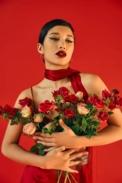 Youthful and sensual asian woman with bold makeup and brunette hair embracing bouquet of roses while posing in neckerchief on red background, fashionable spring, generation z — Stock Photo