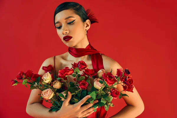 Alluring and young asian fashion model in stylish neckerchief, with brunette hair and bold makeup hugging bouquet of fresh roses on red background, spring fashion photography — Stock Photo