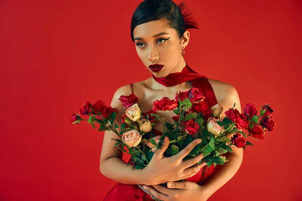 Elegant asian fashion model with bold makeup, brunette hair and expressive gaze holding bouquet of roses and looking away on red background, generation z, trendy spring — Stock Photo