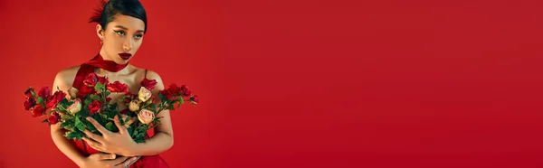 Attraente donna asiatica con capelli castani e trucco audace, indossa un brillante fazzoletto, tiene bouquet di rose fresche e distogliendo lo sguardo su sfondo rosso, concetto di moda primaverile, banner — Foto stock