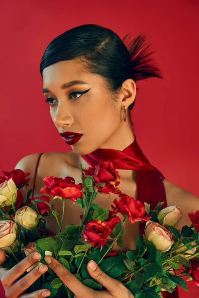 Концепція весняного стилю, молода мода, витончена азіатська жінка з брюнеткою волоссям, сміливий макіяж і модна зачіска, що позує в хустці, тримаючи нові троянди на червоному тлі — стокове фото