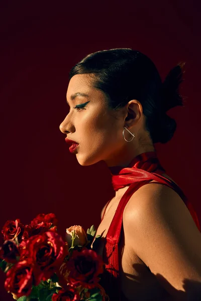 Perfil de mulher asiática encantadora com cabelo morena e maquiagem ousada, em brinco de prata e lenço vermelho segurando buquê de rosas no fundo escuro, fotografia de moda primavera — Fotografia de Stock