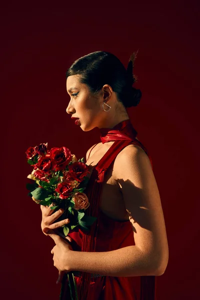 Side view of mesmerizing asian fashion model in red neckerchief, with brunette hair and bold makeup posing with bouquet of roses on dark background, trendy spring, fashion shoot — Stock Photo