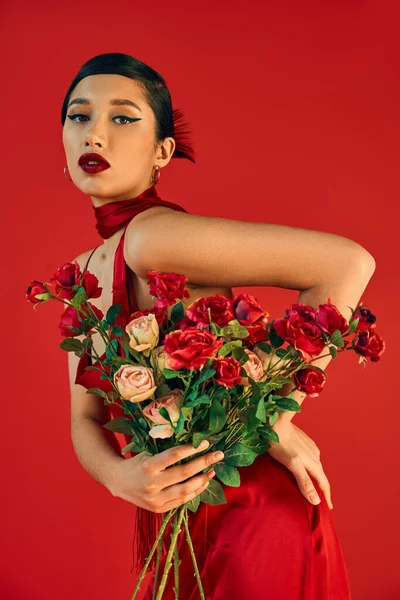 Young and seductive asian woman with bold makeup, in trendy dress posing with hand on hip and roses while looking at camera on red background, trendy spring, fashion shoot — Stock Photo