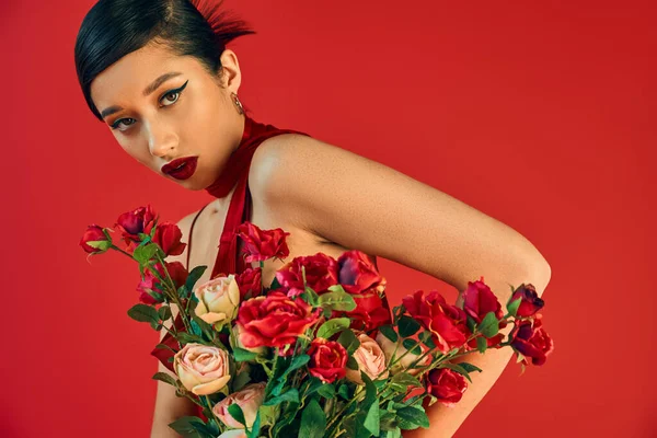 Jovem e hipnotizante mulher asiática com maquiagem ousada, cabelo morena e olhar sedutor para a câmera perto de buquê de rosas frescas no fundo vermelho, primavera na moda, geração z — Fotografia de Stock
