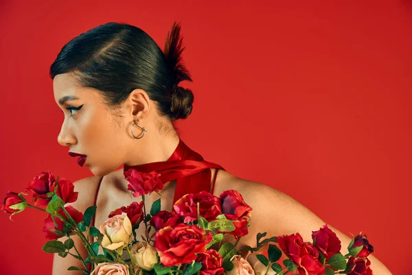 Portrait of beautiful asian woman with brunette hair, bold makeup and trendy hairstyle, wearing neckerchief and looking away near bouquet of roses on red background, trendy spring concept — Stock Photo
