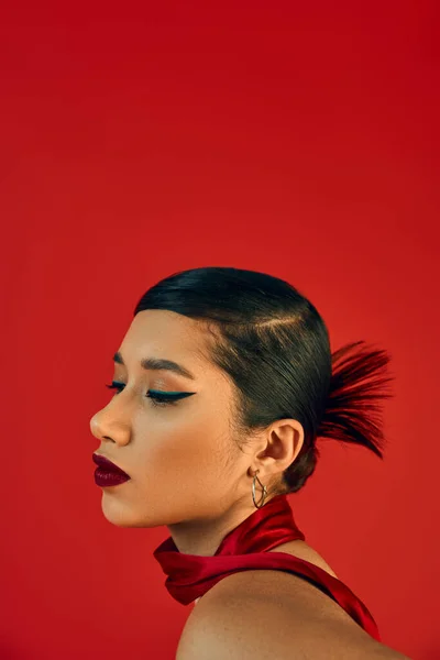 Moda juvenil, elegante concepto de primavera, retrato de mujer asiática joven y encantadora con cabello moreno, maquillaje audaz y peinado de moda posando en el pañuelo sobre fondo rojo - foto de stock