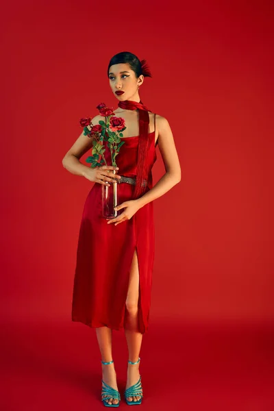 Полная длина молодой и элегантной азиатской женщины в стильном платье ремешок и шейный платок, с смелым макияжем и брюнетки волосы стоят с розами на красном фоне, модный весной, поколение z — стоковое фото