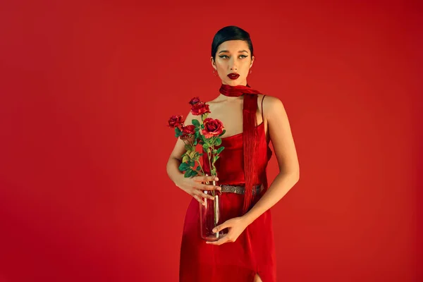 Мода, стильная весенняя концепция, очаровательная женщина в элегантном платье и неккершефе, держащая стеклянную вазу с цветами и отводящая взгляд на красном фоне — стоковое фото