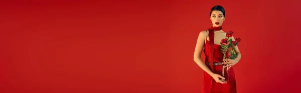Jeune et séduisante femme asiatique avec un maquillage audacieux et cheveux bruns, dans une robe élégante et un foulard poising avec bouquet de roses sur fond rouge, photographie de mode de printemps, bannière — Photo de stock