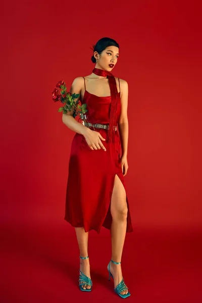 Повна довжина привабливої азіатської жінки у весняному вбранні, що стоїть з квітами на червоному тлі, елегантна сукня з ремінцем, носовичок, бірюзові сандалі, модна стрічка — стокове фото