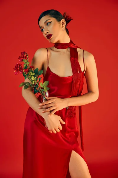 Primavera de moda, moda juvenil, atractiva mujer asiática con cabello moreno y maquillaje audaz, en elegante vestido de correa y pañuelo sosteniendo jarrón de vidrio con rosas frescas sobre fondo rojo - foto de stock