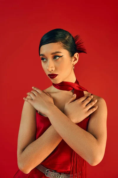 Youthful and elegant asian woman in neckerchief and dress, with brunette hair and bold makeup holding hands on chest and looking away on red background, spring fashion, generation z — Stock Photo