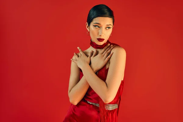 Young asian woman with brunette hair and bold makeup holding hands on chest and looking away while posing in neckerchief and dress on red background, fashionable spring — Stock Photo