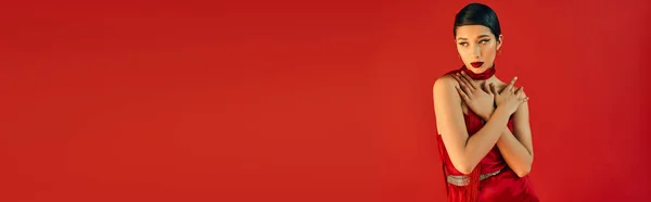 Bela e jovem mulher asiática em vestido elegante e pescoço, com cabelo morena e maquiagem ousada, de mãos dadas perto do rosto e olhando para o fundo vermelho, gen z, moda primavera, banner — Fotografia de Stock