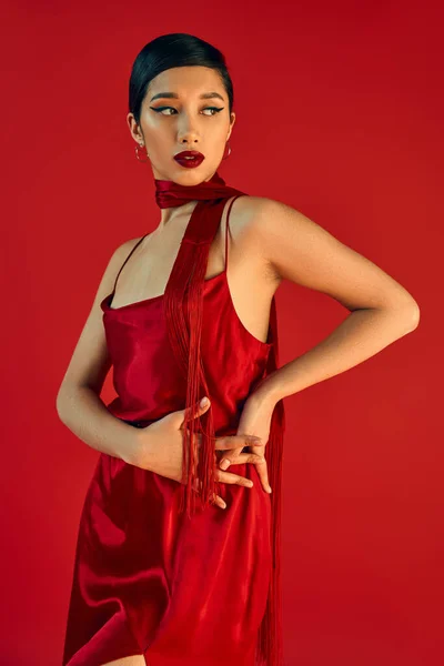 Молода і модна азіатська жінка з сміливим макіяжем і брюнеткою, одягнена в елегантну сукню ремінця і хустку, позуючи руками на стегні на червоному тлі, концепція весняної моди — стокове фото