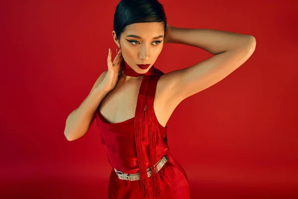 Молодая и выразительная азиатская женщина с брюнетками и смелым макияжем, носящая шейный платок и элегантное платье, отворачивающаяся на красном фоне, ген z мода, стильная весенняя концепция — стоковое фото