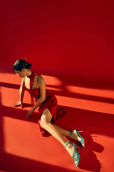 Fotografía de moda de primavera, longitud completa de elegante modelo de moda asiática en vestido de correa, pañuelo y sandalias de color turquesa sentado en la iluminación sobre fondo rojo, vista de ángulo alto - foto de stock