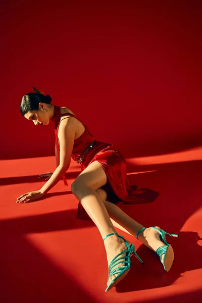 Pleine longueur de femme asiatique expressive et jeune avec des cheveux bruns, dans une robe élégante et des sandales turquoise posant dans l'éclairage sur fond rouge, gen z fashion, concept de style printemps — Photo de stock