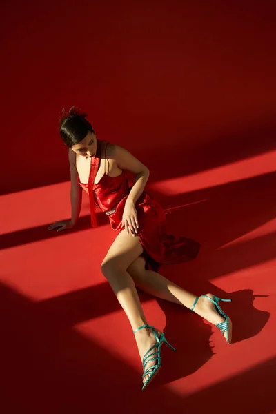 Longitud completa de la mujer asiática de moda en vestido de correa elegante, pañuelo y sandalias de color turquesa sentado sobre fondo rojo con iluminación, vista de ángulo alto, concepto de primavera de moda - foto de stock