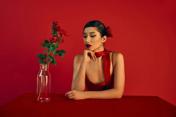 Romántica mujer asiática en vestido y pañuelo, con pelo morena y maquillaje audaz sentado en la mesa jarrón de cristal aseado con rosas sobre fondo rojo, fotografía de moda, elegante concepto de primavera - foto de stock