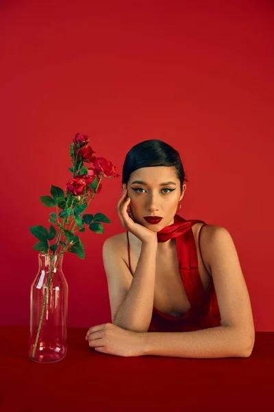 Moda primavera, moda jovem, mulher asiática bonita com maquiagem ousada e cabelo morena sentado no pescoço perto de vaso de vidro com rosas e olhando para a câmera no fundo vermelho — Fotografia de Stock