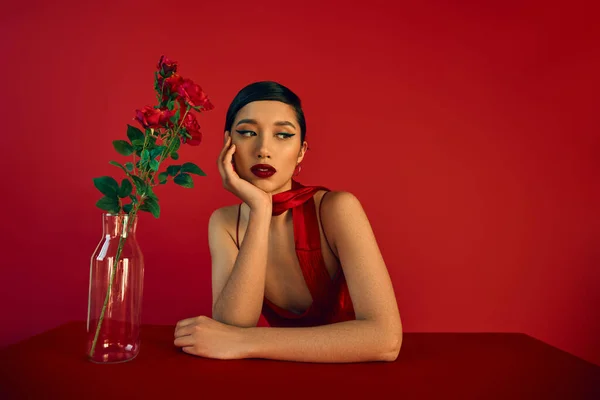 Séduisante femme asiatique aux cheveux bruns et au maquillage audacieux, dans une robe élégante et un foulard assis à table près d'un vase en verre avec des roses fraîches sur fond rouge, concept de mode printanière — Photo de stock