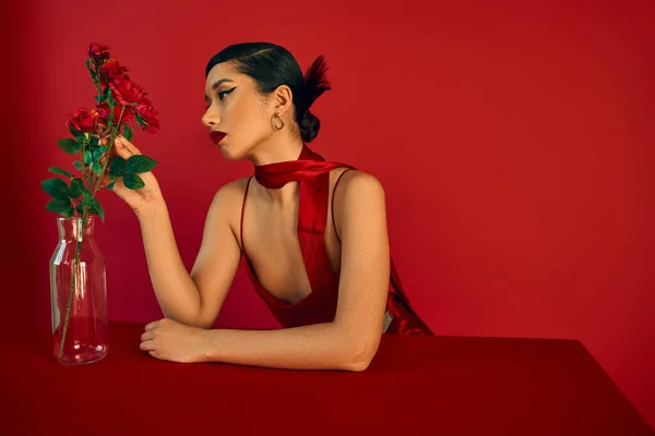 Atemberaubende asiatische Frau in elegantem Trägerkleid, mit brünetten Haaren und kühnem Make-up, das Rosen in einer Glasvase berührt, während sie am Tisch auf rotem Hintergrund sitzt, Frühlingsfotografie — Stockfoto