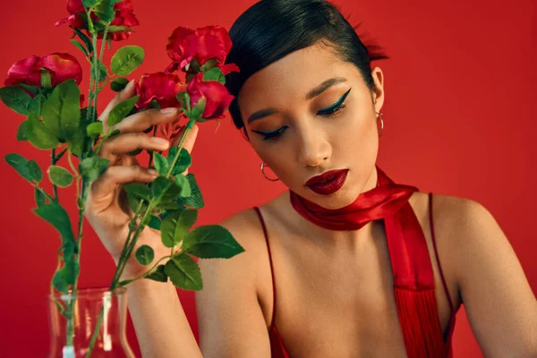 Femme asiatique sensuelle et rêveuse avec maquillage audacieux, cheveux bruns et foulard élégant touchant des roses fraîches sur fond rouge, style printemps, photographie de mode — Photo de stock