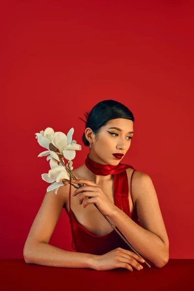 Young and beautiful asian woman with brunette hair and bold makeup holding white orchid while sitting at table in neckerchief and strap dress on red background, trendy spring, gen z fashion — Stock Photo