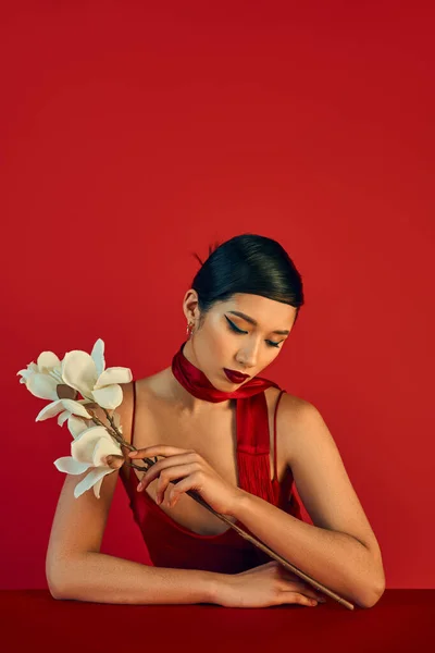 Moda juvenil, concepto de primavera de moda, encantadora mujer asiática en pañuelo y vestido de correa sentado a la mesa con orquídea blanca en flor sobre fondo rojo - foto de stock
