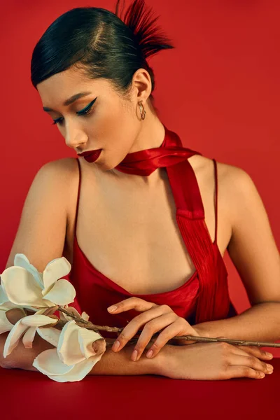 Élégante et jeune femme asiatique en robe tendance et foulard, avec des cheveux bruns et un maquillage audacieux assis à table et regardant orchidée blanche sur fond rouge, gen z fashion, concept printemps tendance — Photo de stock
