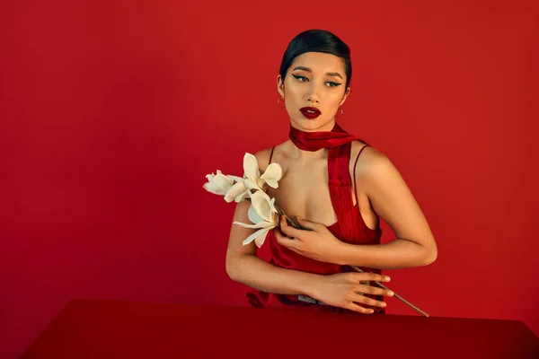 Mujer asiática sensual y morena con maquillaje audaz, en vestido elegante y pañuelo sentado a la mesa con orquídea blanca en flor sobre fondo rojo, generación z, concepto de primavera de moda - foto de stock