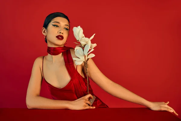 Stylish and charming asian woman in elegant dress and neckerchief, with brunette hair and bold makeup looking at blooming orchid near table on red background, spring fashion photography — Stock Photo