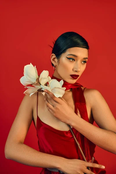 Femme asiatique romantique en tenue de printemps tenant orchidée blanche et regardant loin sur fond rouge, cheveux bruns, maquillage audacieux, robe de sangle, foulard, génération z — Photo de stock