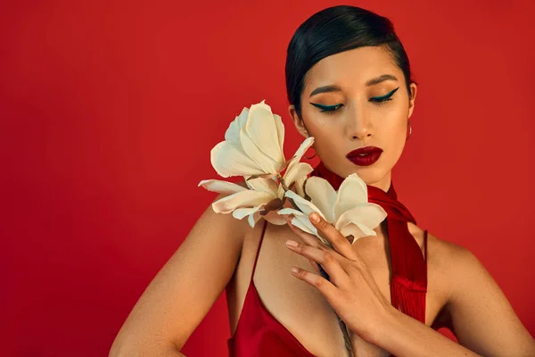 Грациозная и нежная азиатская женщина с брюнетками, смелый макияж, в стильном платье с ремешком и шеей позирует с белой цветущей орхидеей на красном фоне, модная весенняя концепция — стоковое фото