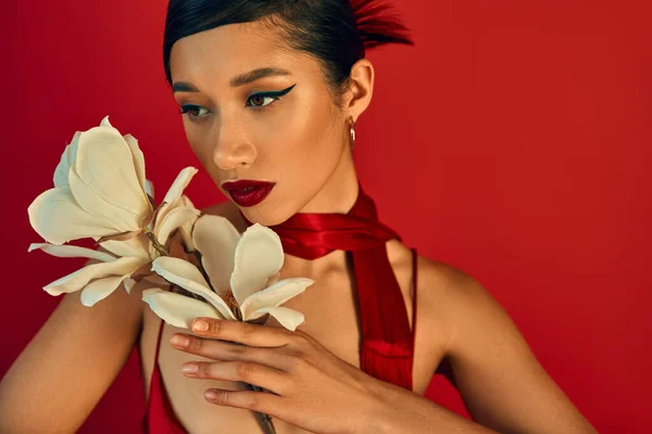Retrato de jovem e atraente mulher asiática com orquídea branca florescendo no fundo vermelho, maquiagem ousada, cabelo morena, lenço de pescoço, fotografia de moda primavera, elegante e charmoso — Fotografia de Stock
