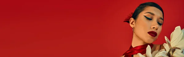 Retrato de hipnotizante mulher asiática com cabelo morena e maquiagem ousada posando com olhos fechados perto de orquídea florescente branca em fundo vermelho, fotografia de moda primavera, banner — Fotografia de Stock