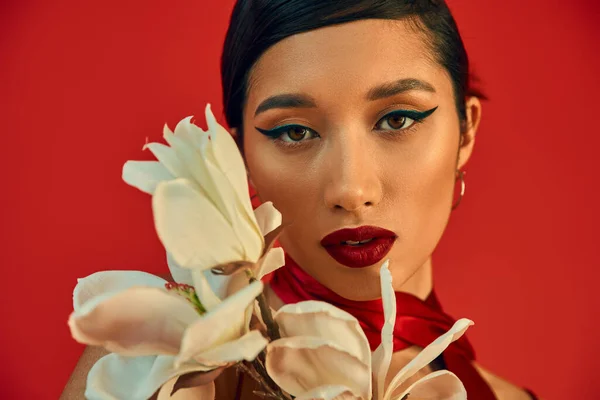Retrato de mujer asiática joven y fascinante con cabello moreno, maquillaje audaz y mirada expresiva mirando a la cámara cerca mientras florece orquídea sobre fondo rojo, fotografía de moda de primavera - foto de stock
