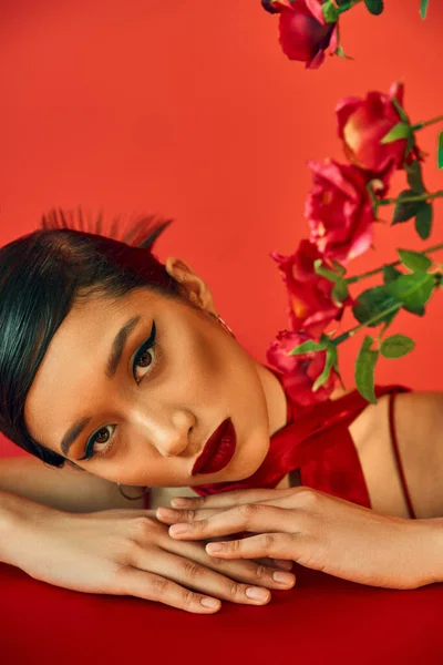 Портрет молодой и привлекательной азиатской модели с смелым макияжем и брюнеткой, смотрящей в камеру, лежащей на столе рядом с розами на красном фоне, стильной весной, модной съемкой — стоковое фото