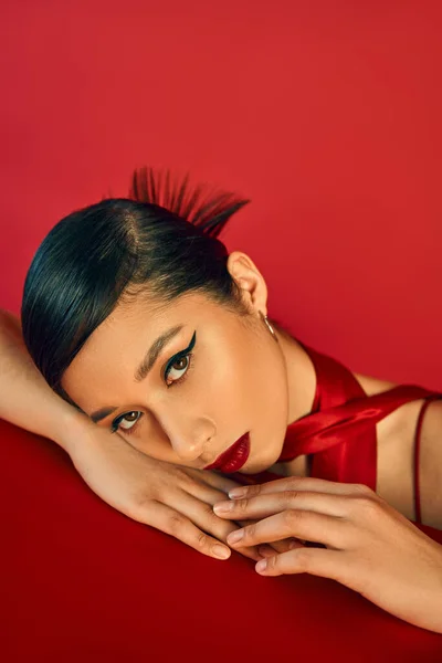 Spring fashion concept, portrait of young and beautiful asian fashion model leaning on table and looking at camera on red background, brunette hair, bold makeup, expressive gaze, generation z — Stock Photo