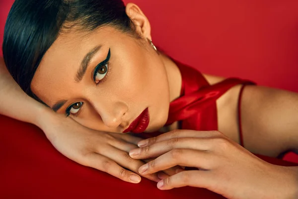 Портрет молодой и чувственной азиатской женщины с смелым макияжем и брюнеткой, лежащей на столе и смотрящей на камеру на красном фоне, поколение z, весенняя модная фотография — стоковое фото