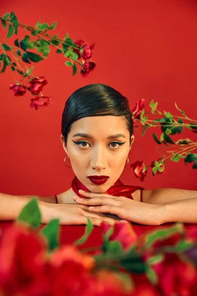 Portrait de jeune et charmante femme asiatique avec maquillage lumineux et cheveux bruns regardant la caméra entourée de roses fraîches sur fond rouge, concept de photographie de mode de printemps — Photo de stock