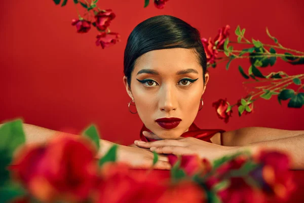 Gen z fashion, портрет очаровательной и молодой азиатки с ярким макияжем, смотрящей в камеру среди свежих роз на красном, весенняя модная фотография, размытый передний план — стоковое фото