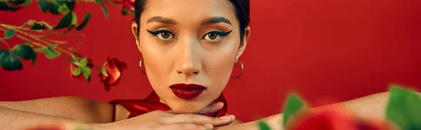 Портрет молодой и очаровательной азиатской модели с ярким макияжем и выразительным взглядом на камеру рядом с цветами на красном фоне, модная весенняя концепция, баннер — стоковое фото