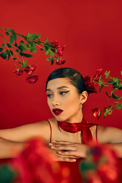 Retrato de mujer asiática elegante y hermosa con maquillaje audaz, cabello moreno y pañuelo apoyado en la mesa y mirando hacia otro lado cerca de rosas en rojo, borrosa primer plano - foto de stock