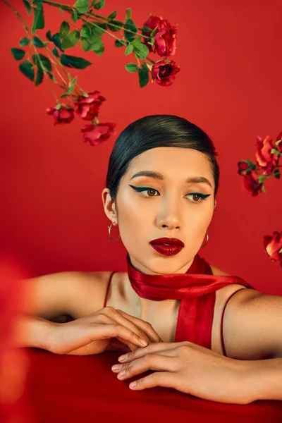 Retrato de fascinante y soñadora mujer asiática posando cerca de rosas frescas sobre fondo rojo, maquillaje audaz, cabello moreno, elegante pañuelo, concepto de primavera de moda — Stock Photo