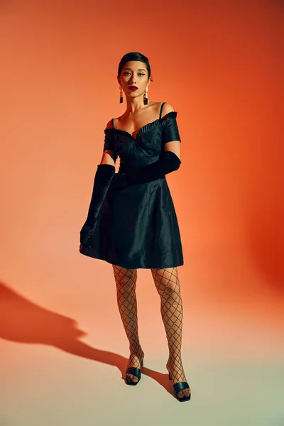 Повна довжина молодої і елегантної азіатки в чорній коктейльній сукні, довгі рукавички і ажурні колготки стоять і дивляться на камеру на червоному і помаранчевому фоні, концепція весняної моди — стокове фото