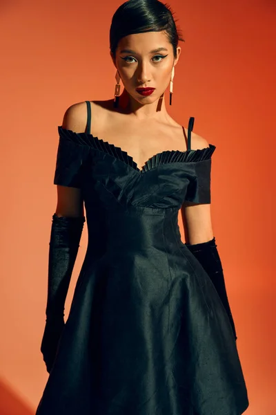 Longitud completa de la mujer asiática de moda y joven en guantes, vestido de cóctel negro y pendientes de moda mirando a la cámara con mirada seductora sobre fondo rojo y naranja - foto de stock